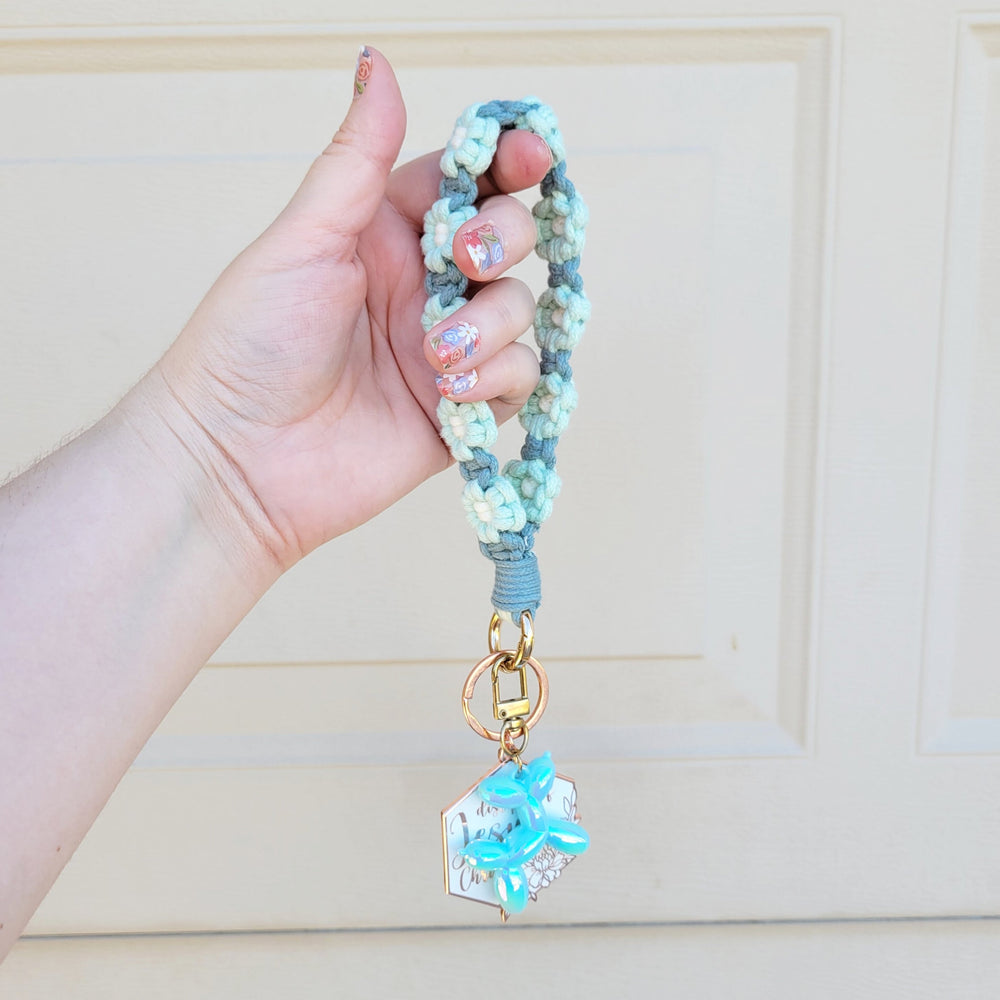 Teal and Blue Flower Macrame Wristlet Keychain with gold clip, being held in one hand, hanging in the air with several keychains attached