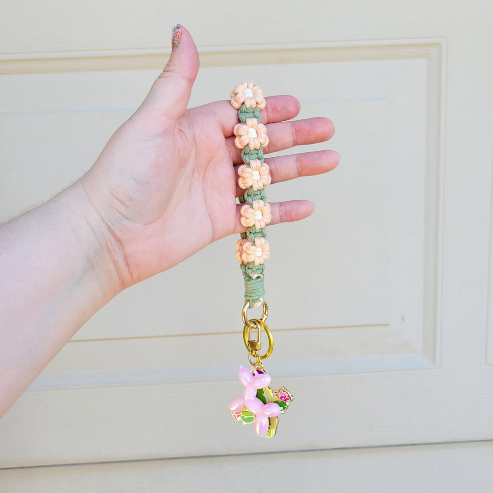 Sage and Pink Flower Macrame Wristlet Keychain with gold clip, being held in one hand, hanging in the air with several keychains attached