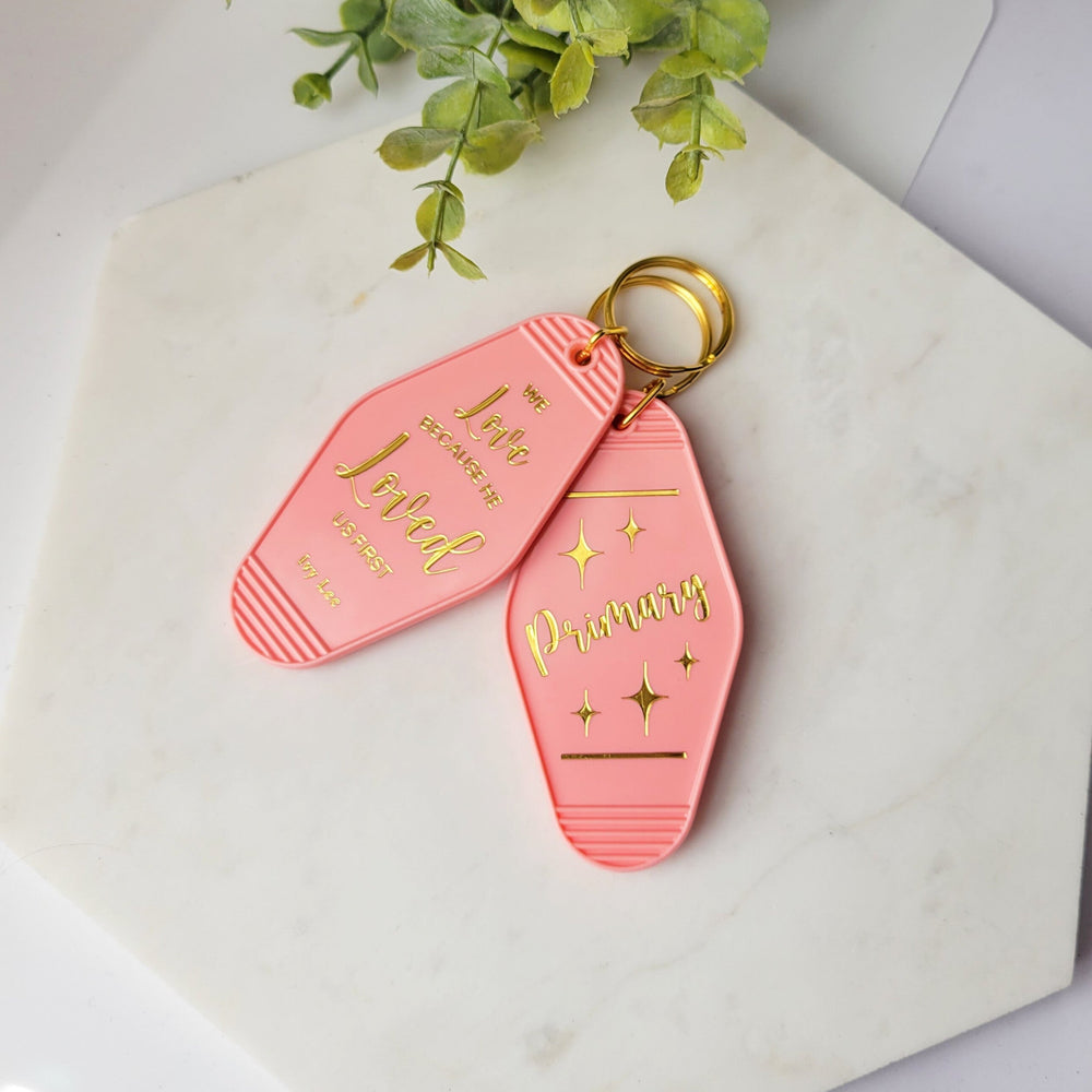 retro shape salmon pink keychain, with 'Primary' in gold foil, and a second keychains with 'We love Because He loved us first" in gold foil
