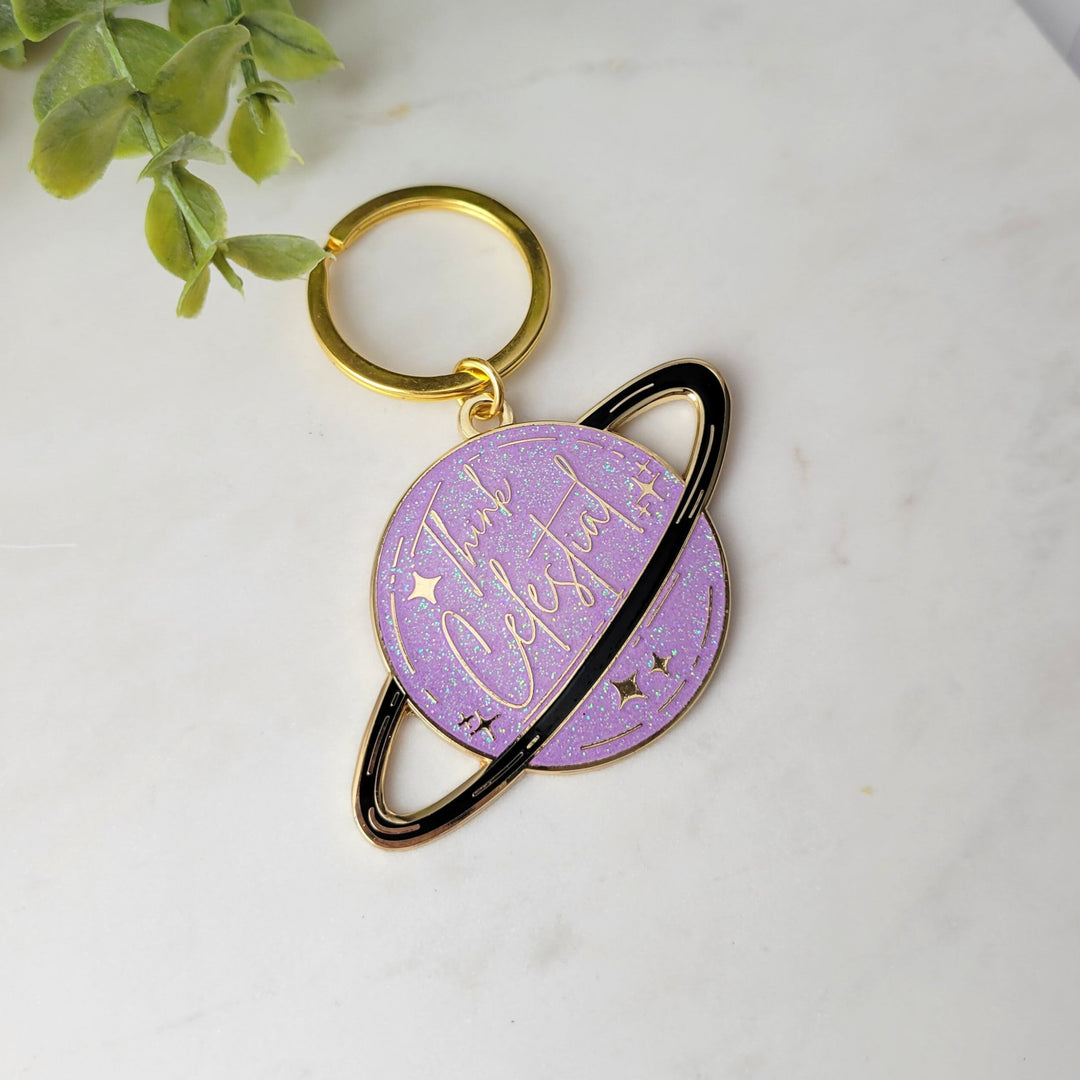 A planet shaped purple keychain, with a black ring around the planet, with stars and glitter, and Think celestial lettering, with a gold plating on tile