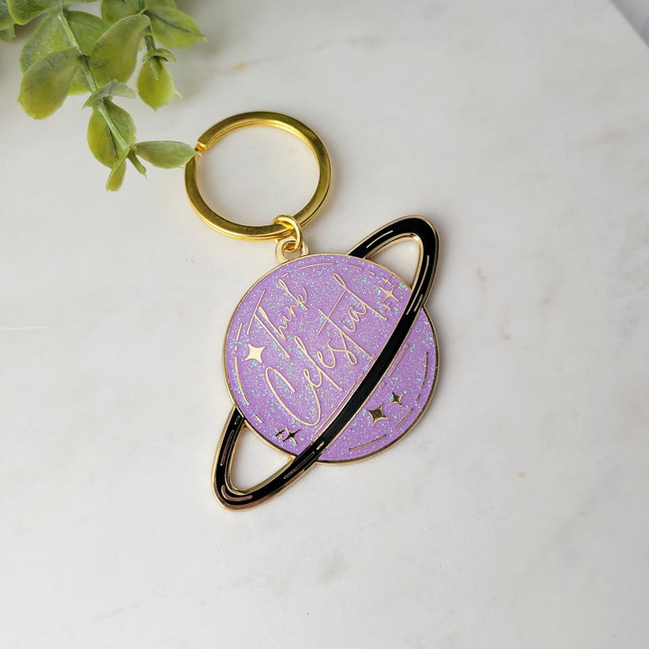 A planet shaped purple keychain, with a black ring around the planet, with stars and glitter, and Think celestial lettering, with a gold plating on tile