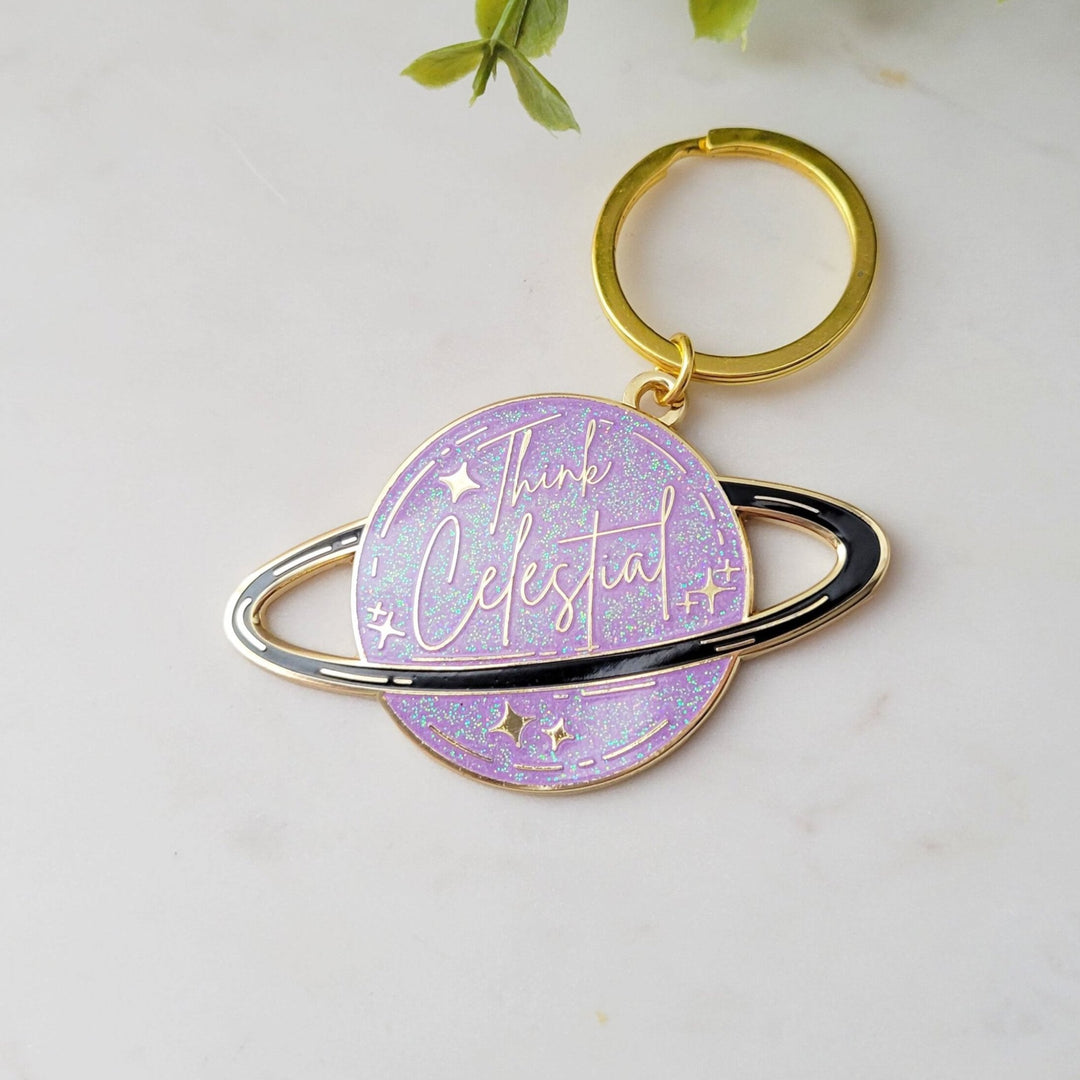 A planet shaped purple keychain, with a black ring around the planet, with stars and glitter, and Think celestial lettering, with a gold plating