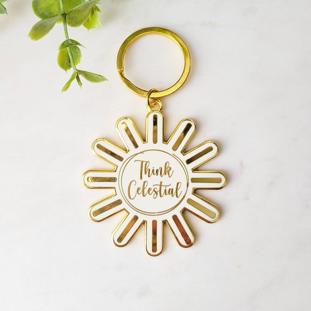 A white sun shaped keychain, with Think celestial lettering, and gold plating and ring