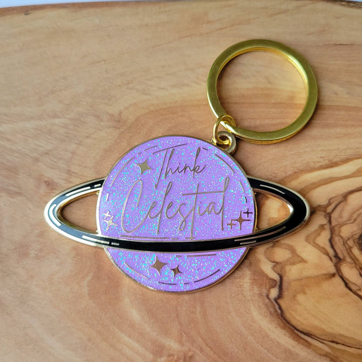 A planet shaped purple keychain, with a black ring around the planet, with stars and glitter, and Think celestial lettering, with a gold plating, close up on wood