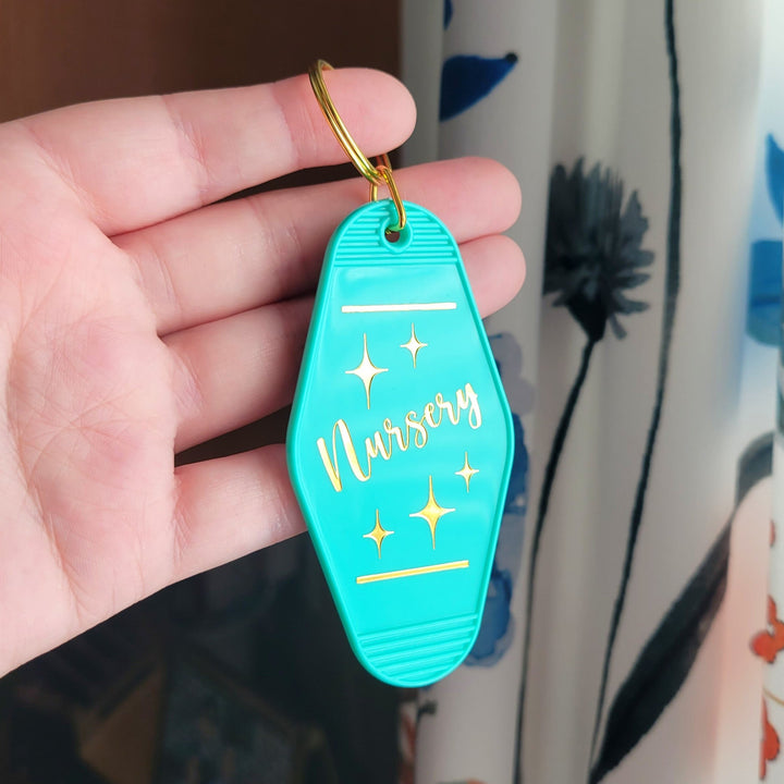 teal colored retro keychain with the words 'Nursery' in gold foil, being held up