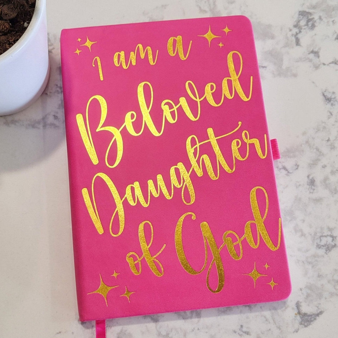 a hot pink colored dot grid journal with gold foil lettering, of I am a beloved daughter of god, laying on a marble counter