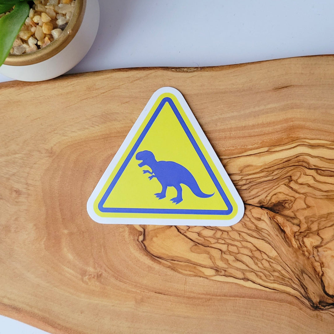 A sticker of a yellow triangle caution sign, with a blue dinosaur t-rex in the middle.