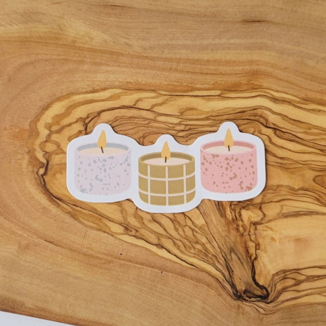 Three small candles in a row, with fires lit. One is blue and tan, the middle is tan and olive colored, and the third is pink and olive.