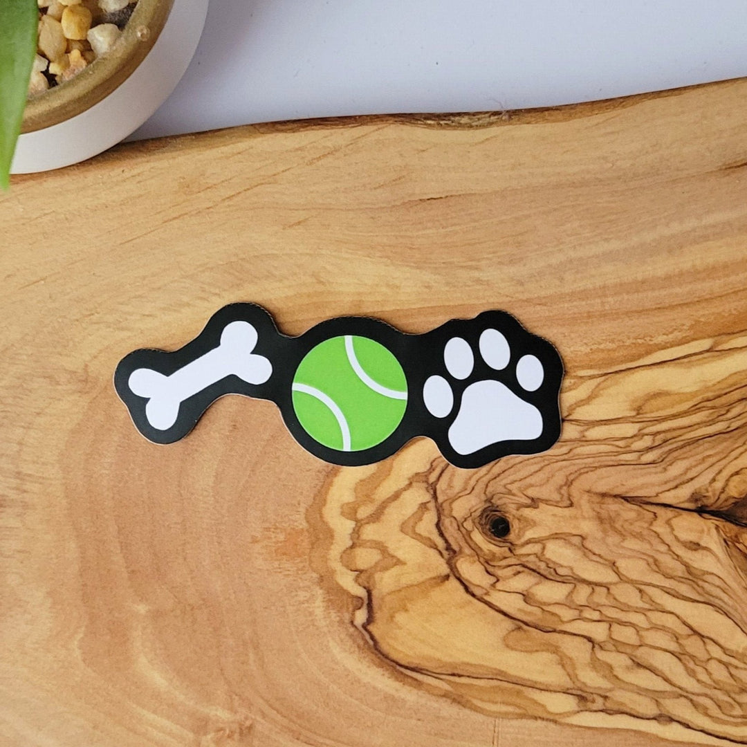 a sticker with three items in a row, a white bone, green tennis ball, and a white paw print, all on a black background