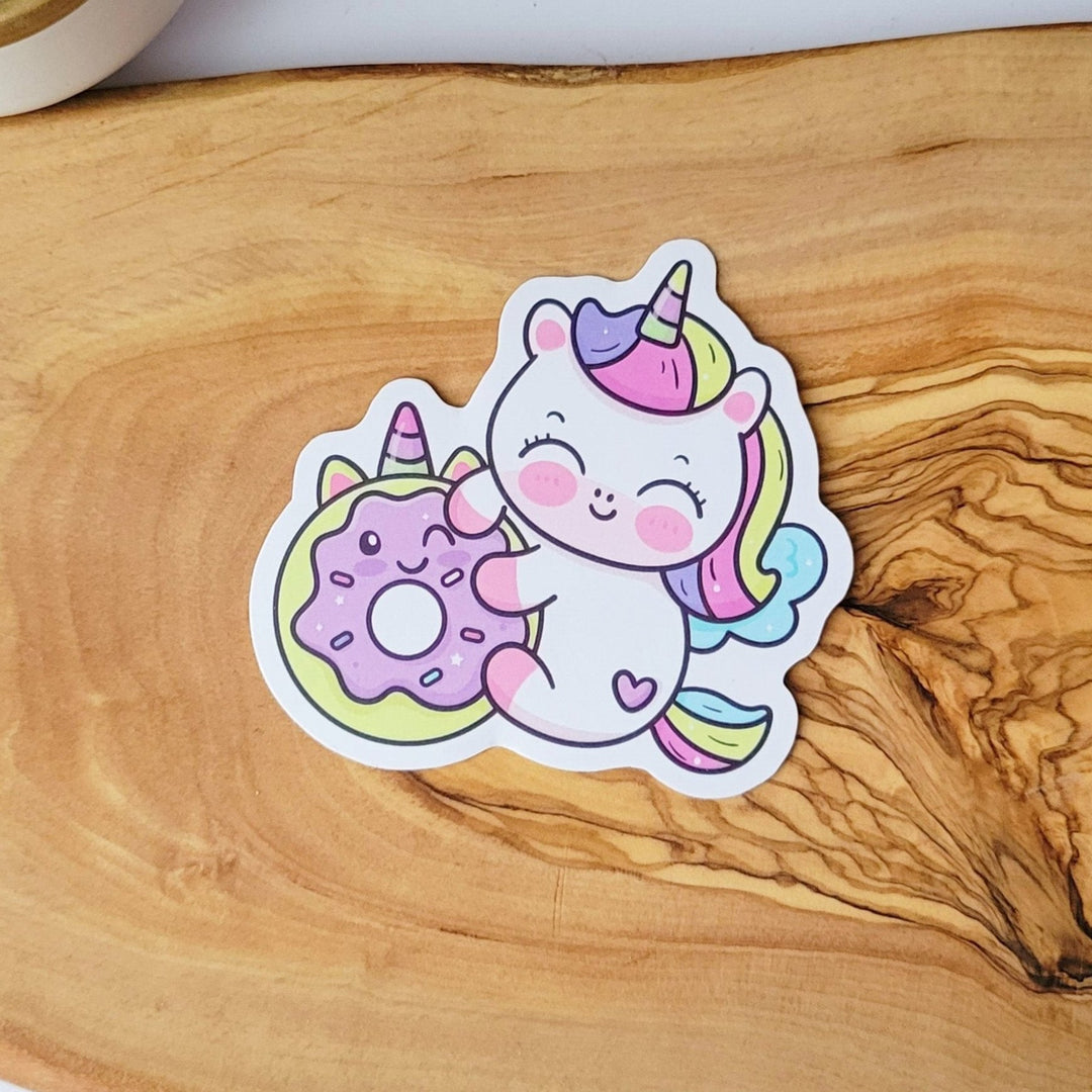a white unicorn with rainbow colored hair, is hugging a purple donut