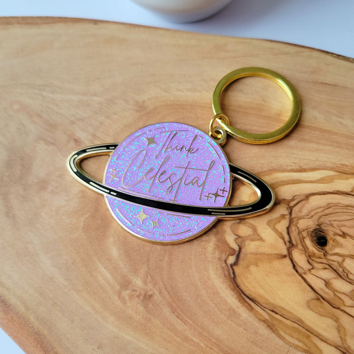 A planet shaped purple keychain, with a black ring around the planet, with stars and glitter, and Think celestial lettering, with a gold plating on wood