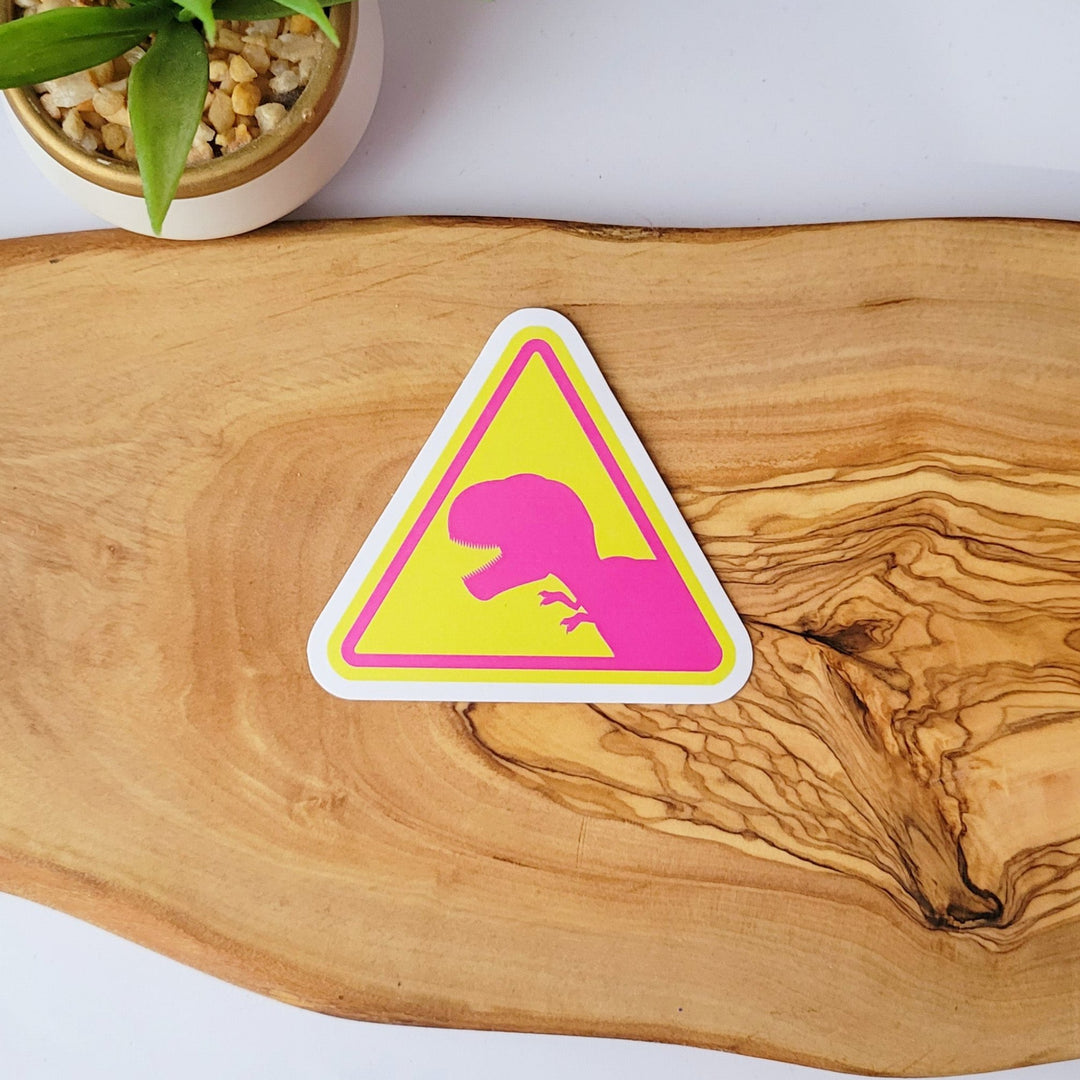 A sticker of a yellow triangle caution sign, with a pink dinosaur t-rex in the middle.
