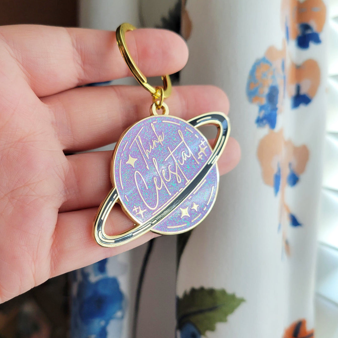 A planet shaped purple keychain, with a black ring around the planet, with stars and glitter, and Think celestial lettering, with a gold plating being held up by a hand