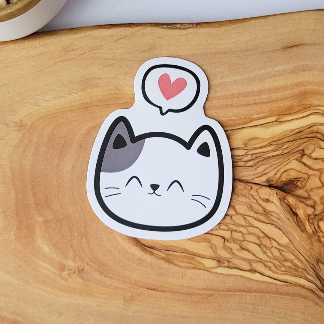 a cute sticker of a smiling happy white cat face, with a grey ear patch, with a conversation bubble with a heart in it, above the cat face.