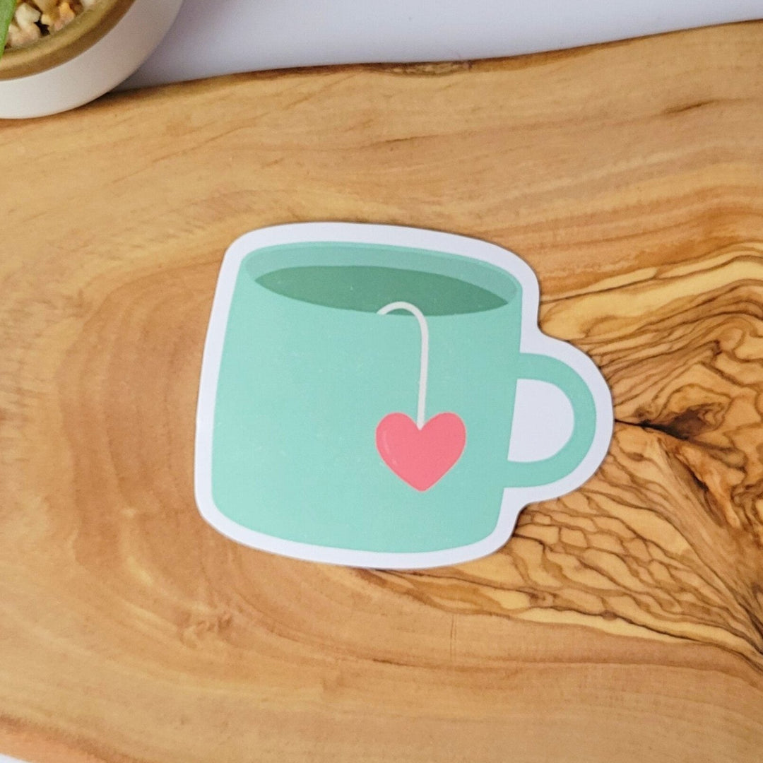 a green cup with a tea bag handle in the shape of a heart, lying over the side of the mug