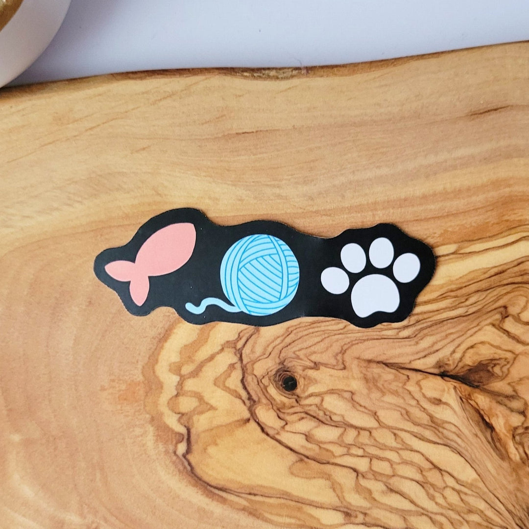A small sticker with of three items in a line, a salmon colored fish, a blue ball of yarn, and a white cat paw, all on a black background.