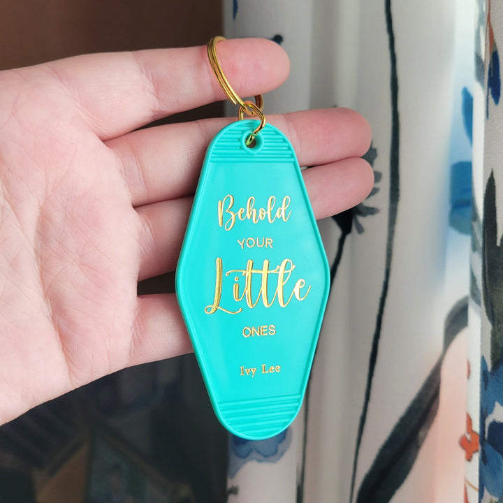 teal colored retro keychain with the words 'Behold your little ones' in gold foil, being held up