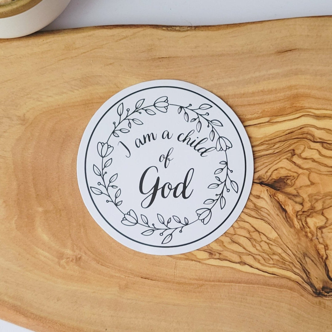 A white circle sticker with I am a child of God lettering, with a circle wreath of calm leaves and flowers.