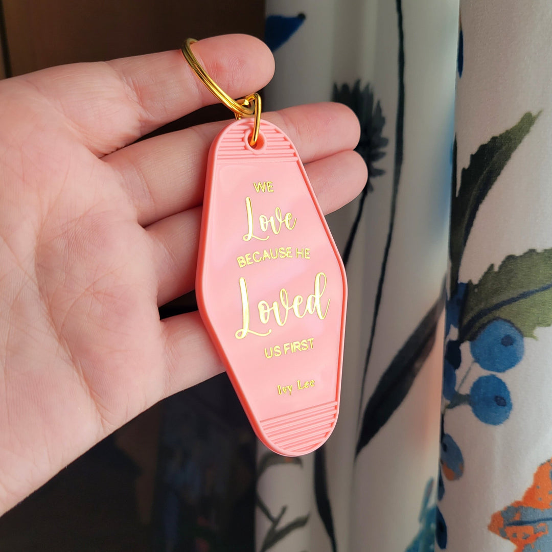 salmon pink keychain with 'We love Because He loved us first" in gold foil, being held up