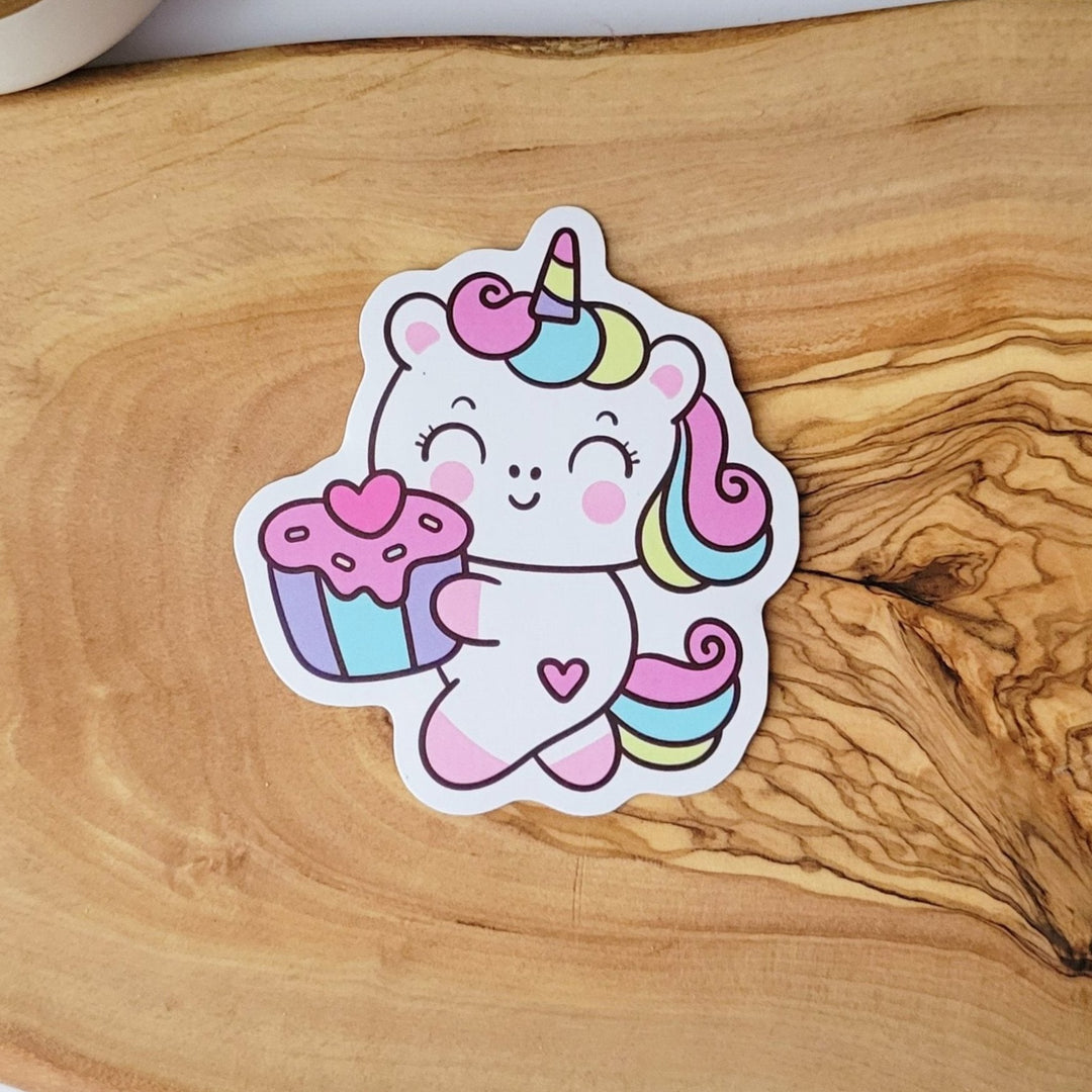 a white unicorn with rainbow colored hair, is holding a pink cupcake