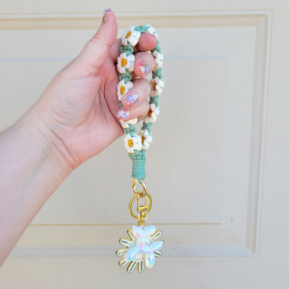 Green and White Flower Macrame Wristlet Keychain with gold clip, being held in one hand, hanging in the air with several keychains attached.