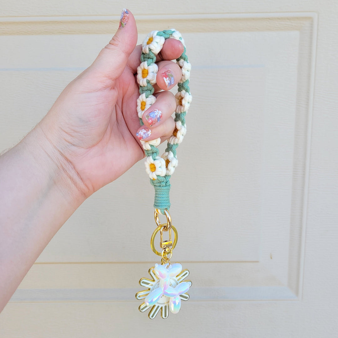 Green and White Flower Macrame Wristlet Keychain with gold clip, being held in one hand, hanging in the air with several keychains attached.