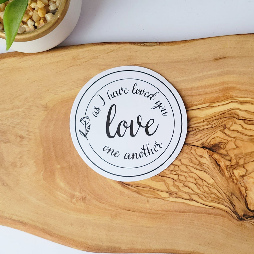 a white circle sticker with the words As I have loved you Love one another, with a simple flower outline