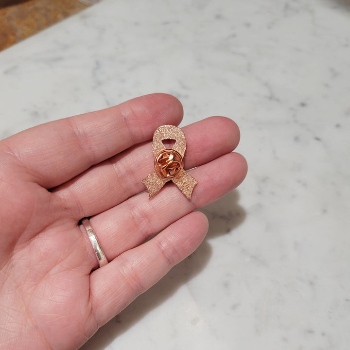 the back of a cancer ribbon shaped pin with copper color clasp