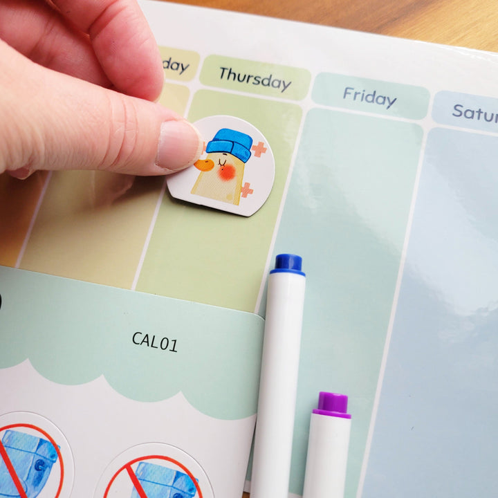 Hand placing a sticker on a planner with a pen next to it